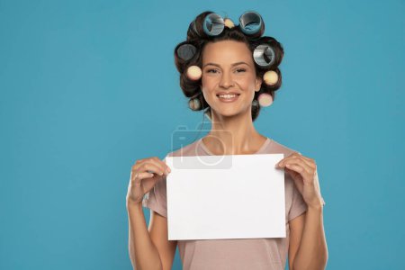 Photo for Beautiful smiling woman with hair curlers holding empty paper on a blue studio  background - Royalty Free Image