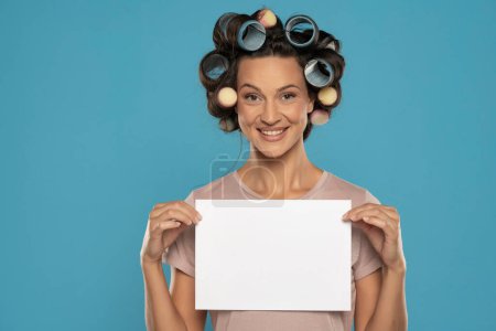 Foto de Beautiful smiling woman with hair curlers holding empty paper on a blue studio  background - Imagen libre de derechos