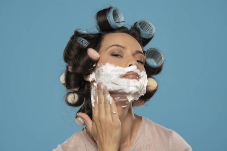 Photo for Attractive young woman with hair curlers rollers applyes shaving foam on her face on a blue studio background - Royalty Free Image