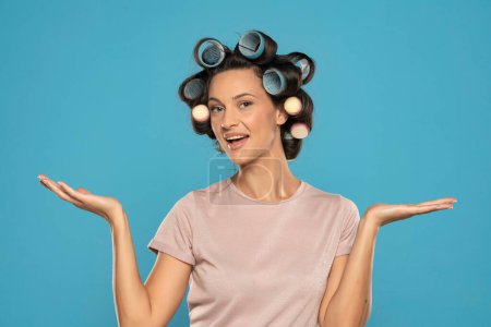 Photo for Beautiful smiling woman with hair curlers advertizing imaginary product on her palms on a blue studio  background - Royalty Free Image