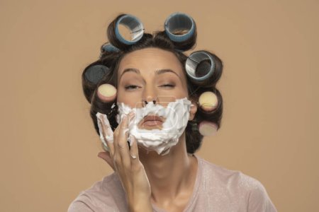 Foto de Attractive young woman with hair curlers rollers applyes shaving foam on her face on a beige studio background - Imagen libre de derechos