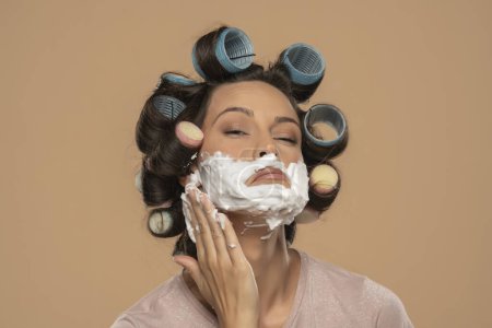 Téléchargez les photos : Attrayant jeune femme avec cheveux bigoudis rouleaux applaudissements mousse à raser sur son visage sur un fond studio beige - en image libre de droit