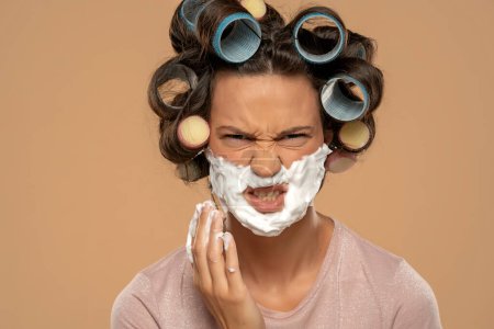 Téléchargez les photos : Attractive angry woman with hair curlers rollers applyes shaving foam on her face on a beige studio background - en image libre de droit