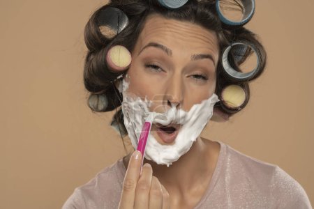 Téléchargez les photos : Attrayant jeune femme avec des boucles de cheveux rouleaux se rase le visage sur un fond de studio beige - en image libre de droit