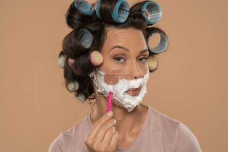 Foto de Atractiva mujer joven con rulos de pelo rodillos se afeita la cara en un fondo de estudio beige - Imagen libre de derechos