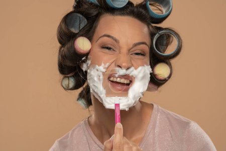Téléchargez les photos : Attractive young funny woman with hair curlers rollers shaves her face on a beige studio background - en image libre de droit