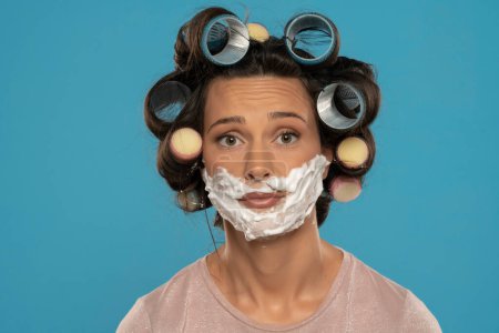 Foto de Atractiva mujer infeliz con rulos de pelo rodillos posando con espuma de afeitar en su cara sobre un fondo de estudio azul - Imagen libre de derechos