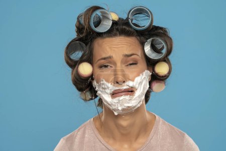 Foto de Atractiva mujer infeliz con rulos de pelo rodillos posando con espuma de afeitar en su cara sobre un fondo de estudio azul - Imagen libre de derechos