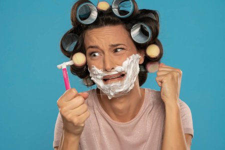 Téléchargez les photos : Attractive desperate woman with hair curlers rollers posing with shaving foam and razor blade on a blue studio background - en image libre de droit