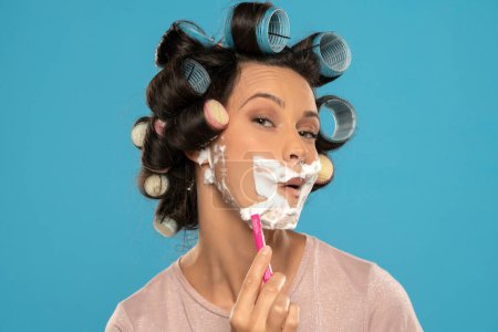 Foto de Atractiva joven con rizadores de pelo se afeita la cara en un fondo de estudio azul - Imagen libre de derechos