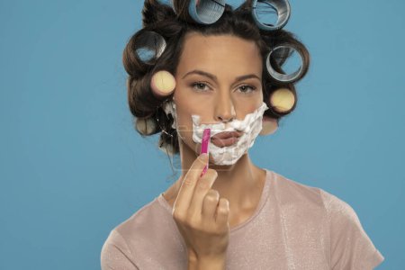 Téléchargez les photos : Attractive young woman with hair curlers shaves her face on a blue studio background - en image libre de droit