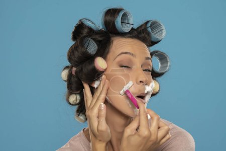 Foto de Atractiva joven divertida mujer con rulos de pelo rodillos se afeita la cara en un fondo de estudio azul - Imagen libre de derechos