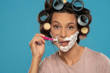 Foto de Atractiva joven divertida mujer con rulos de pelo rodillos se afeita la cara en un fondo de estudio azul - Imagen libre de derechos