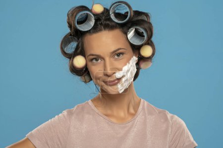 Téléchargez les photos : Attractive happy  woman with hair curlers rollers posing with shaving foam on her face on a blue studio background - en image libre de droit