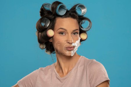 Téléchargez les photos : Séduisante femme avec des boucles de cheveux rouleaux posant avec de la mousse à raser sur son visage sur un fond bleu studio - en image libre de droit