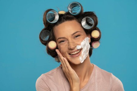 Téléchargez les photos : Séduisante femme heureuse avec des boucles de cheveux rouleaux posant avec de la mousse à raser sur son visage sur un fond bleu studio - en image libre de droit