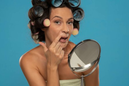 Téléchargez les photos : Femme avec des bigoudis applique correcteur sur ses lèvres supérieures sur un fond de studio bleu - en image libre de droit