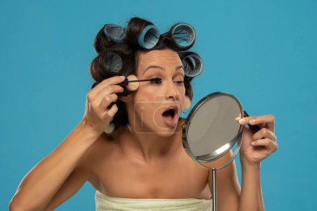 Foto de Mujer con rizadores de pelo aplica rímel en sus pestañas sobre un fondo de estudio azul - Imagen libre de derechos