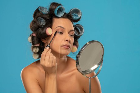 Téléchargez les photos : Femme avec des bigoudis applique mascara sur ses cils sur un fond bleu studio - en image libre de droit