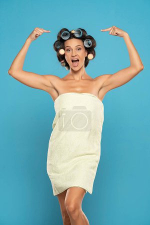 Téléchargez les photos : Young happy woman with hair curlers and towel posing in a blue studio background - en image libre de droit