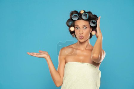 Téléchargez les photos : Jeune femme exscitée avec boucles de cheveux et serviette sur fond bleu isolé tenant copyspace imaginaire sur la paume pour insérer une annonce - en image libre de droit