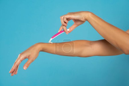Téléchargez les photos : Une femme se rase les bras avec un rasoir rose jetable sur fond de studio bleu - en image libre de droit