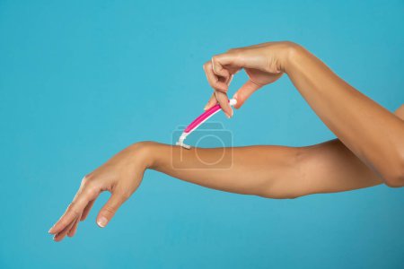 Téléchargez les photos : A woman shaves her arms with a disposable pink razor on a blue studio background - en image libre de droit