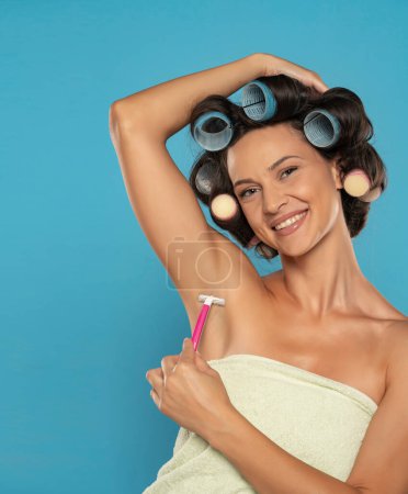 Téléchargez les photos : Une femme heureuse avec des bigoudis se rase les aisselles avec un rasoir rose jetable sur un fond de studio bleu - en image libre de droit
