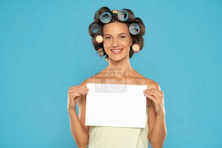 Téléchargez les photos : Beautiful smiling woman with hair curlers holding empty paper on a blue studio  background - en image libre de droit