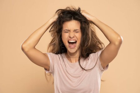 Photo for Angry nervous woman pulling her messy long hair on a beige studio background - Royalty Free Image