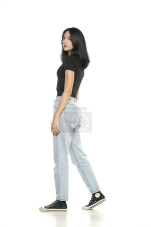 Photo for Side view of a young woman in loose jeans walking on white studio background - Royalty Free Image