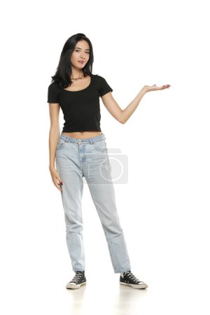 Téléchargez les photos : Jeune femme en jeans et chemise, produit publicitaire sur sa paume, vue de face sur fond de studio blanc - en image libre de droit
