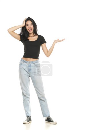 Photo for Young excited woman in jeans and shirt, advetising product on her palm,  front view on a white studio background - Royalty Free Image