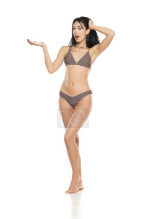 Téléchargez les photos : Jeune femme brune souriante en maillot de bain bikini publicité objet imaginaire sur sa main sur un fond de studio blanc. - en image libre de droit