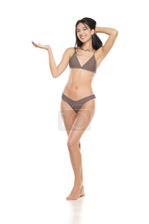 Foto de Joven mujer morena sonriente en bikini traje de baño publicidad objeto imaginario en su mano sobre un fondo de estudio blanco. - Imagen libre de derechos