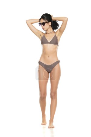 Téléchargez les photos : Jeune femme brune en maillot de bain bikini et lunettes de soleil posant sur un fond de studio blanc. - en image libre de droit