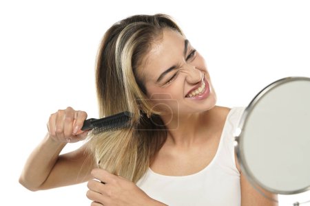 Téléchargez les photos : Jeune femme nerveuse malheureuse se brossant les cheveux devant un miroir sur fond de studio blanc - en image libre de droit