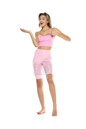 Téléchargez les photos : Femme souriante dans des leggings roses et en haut présentant quelque chose sur sa paume vide sur un fond de studio blanc - en image libre de droit