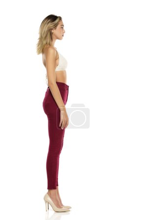 Photo for A young woman in burgundy jeans and top, and high heels shoes posing on a white studio background. Side profile view - Royalty Free Image