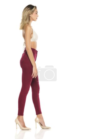 Téléchargez les photos : Une jeune femme en jean bordeaux et haut, et des chaussures à talons hauts posant sur un fond de studio blanc. Vue de profil latérale - en image libre de droit