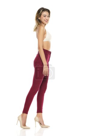 Photo for A young smiling woman in burgundy jeans and top, and high heels shoes posing on a white studio background. Side profile view - Royalty Free Image