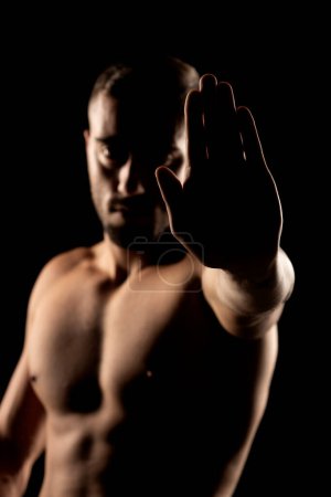 Photo for Silhouette of a muscular shirtless man in the shadow showing stop hand on a black studio background - Royalty Free Image