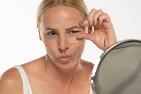 Téléchargez les photos : Portrait d'une femme âgée sans maquillage pincant les rides de ses yeux devant un miroir sur fond de studio blanc - en image libre de droit