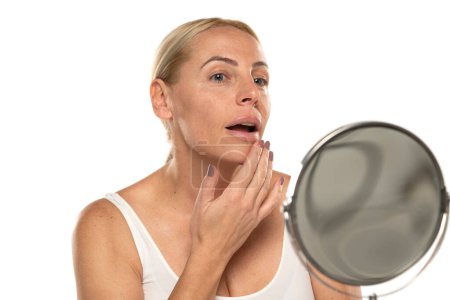 Téléchargez les photos : Inquiet femme blonde d'âge moyen devant le miroir touche sa bouche sur fond de studio blanc. - en image libre de droit