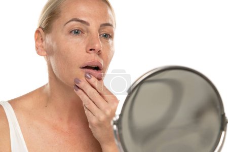 Téléchargez les photos : Inquiet femme blonde d'âge moyen devant le miroir touche sa bouche sur fond de studio blanc. - en image libre de droit
