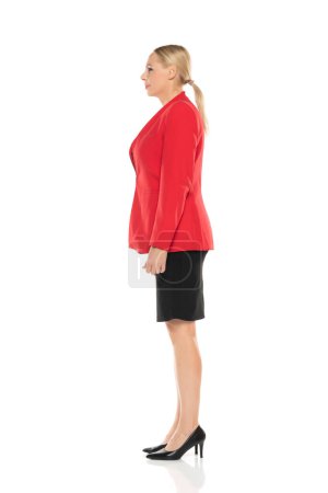 Téléchargez les photos : Femme d'affaires senior d'âge moyen en veste rouge et jupe noire posant sur fond de studio blanc. Côté, vue de profil. - en image libre de droit