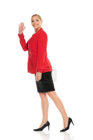Photo for Middle aged senior smiling business woman in red jacket and black skirt walking on white studio background and wave hand. Side, profile view. - Royalty Free Image