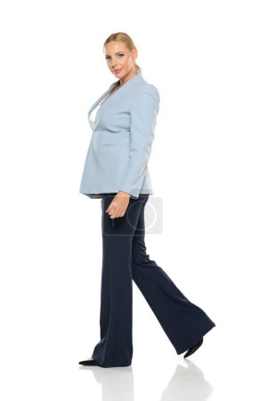 Photo for Middle aged senior smiling woman  wearing blue jacket and black trousers walking on a white studio background. Profile view - Royalty Free Image