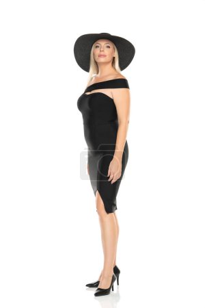 Téléchargez les photos : Femme âgée d'âge moyen en robe noire serrée et chapeau posant sur un fond de studio blanc - en image libre de droit