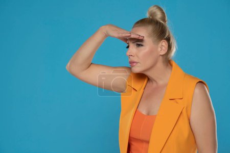 Photo for Curious blonde woman inyellow sleeveless jacket looking far away with hand over head, trying to see something, bad vision. Indoor studio shot isolated on blue background - Royalty Free Image